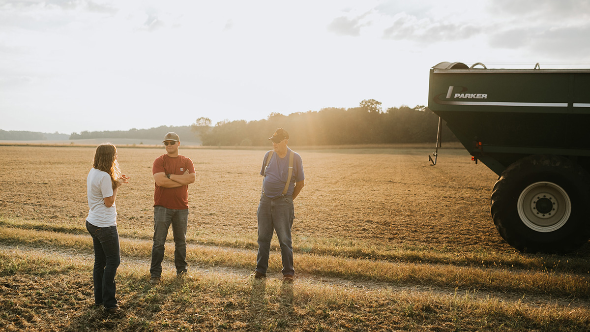 Farmers talking in soybean field during harvest with farm equipment in the background.