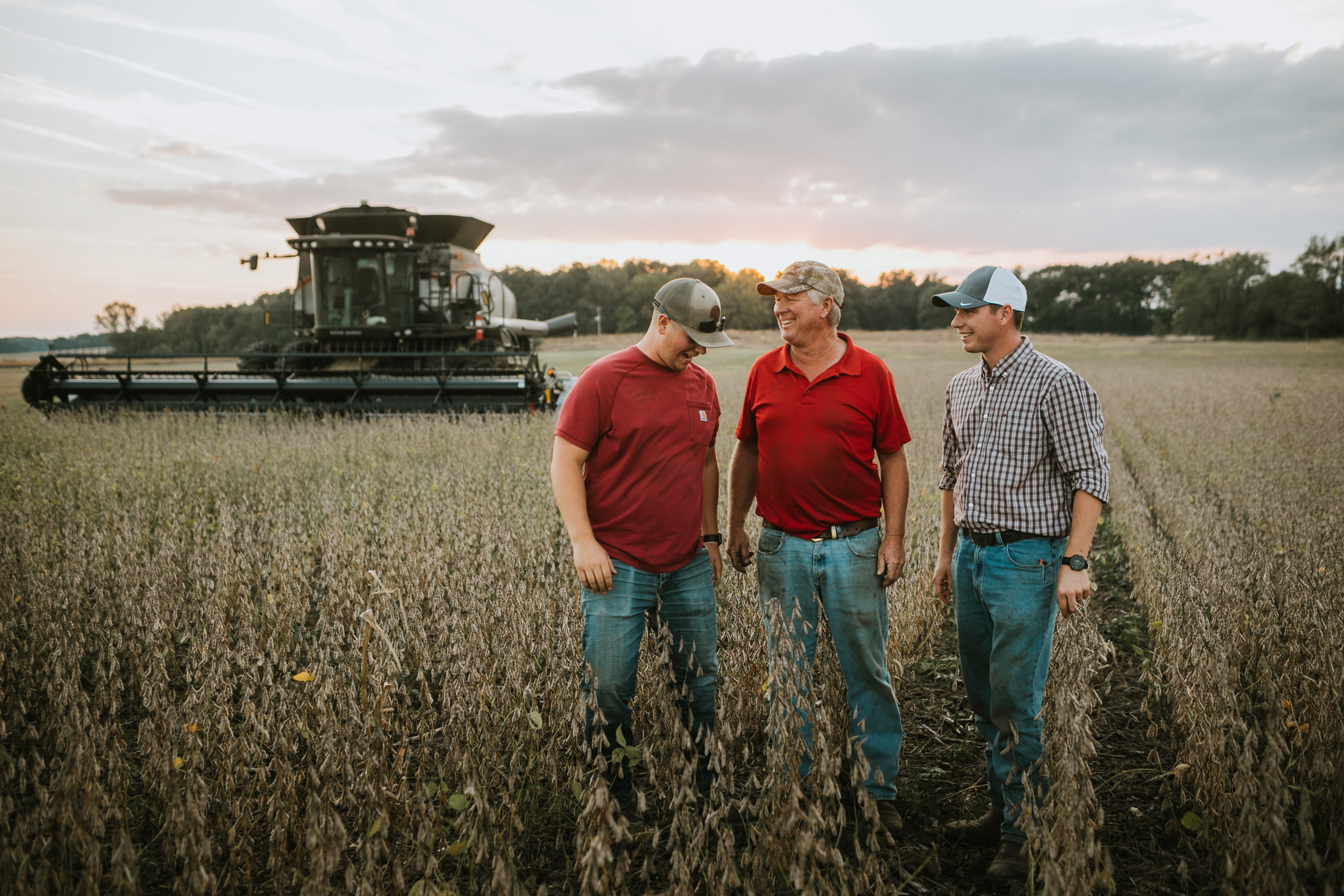 Photo of two generations of farmers standing in a field with a combine in the background.