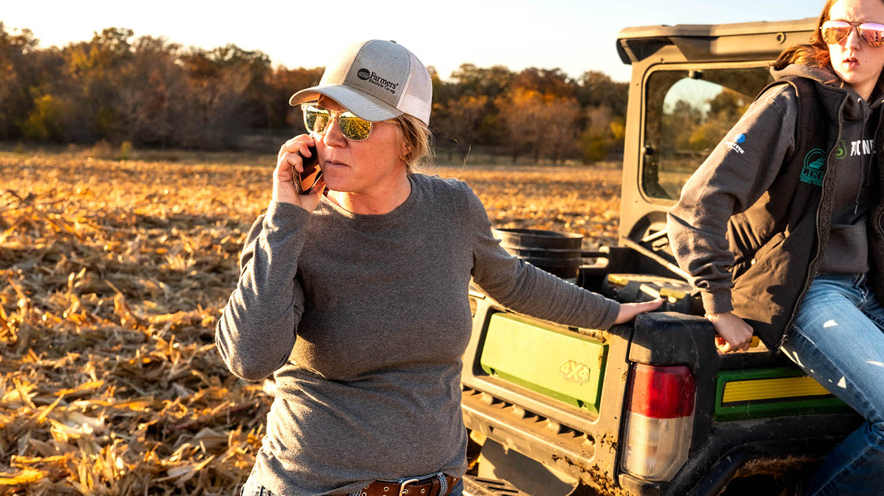 A female farmer talks on the phone in the field while another female sits on the back of a side-by-side vehicle nearby.