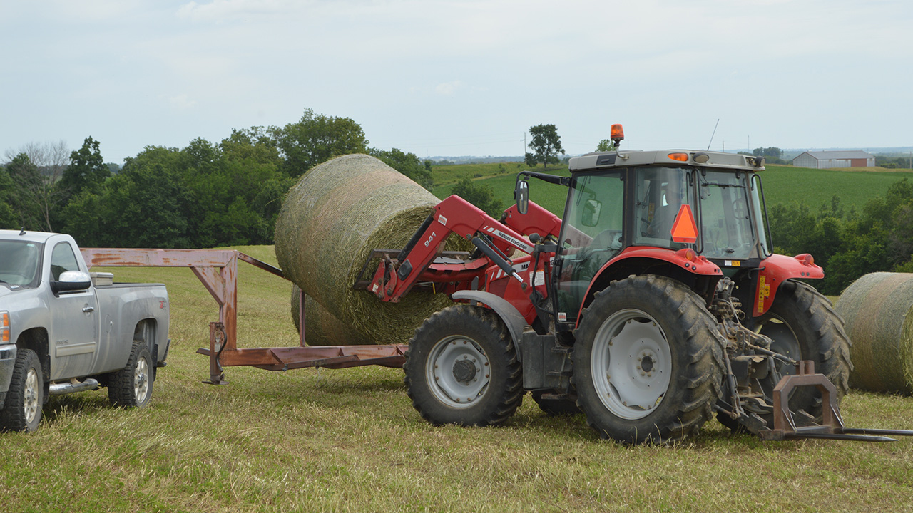 Red tractor lifting a hay bale near a silver truck in a grassy field.