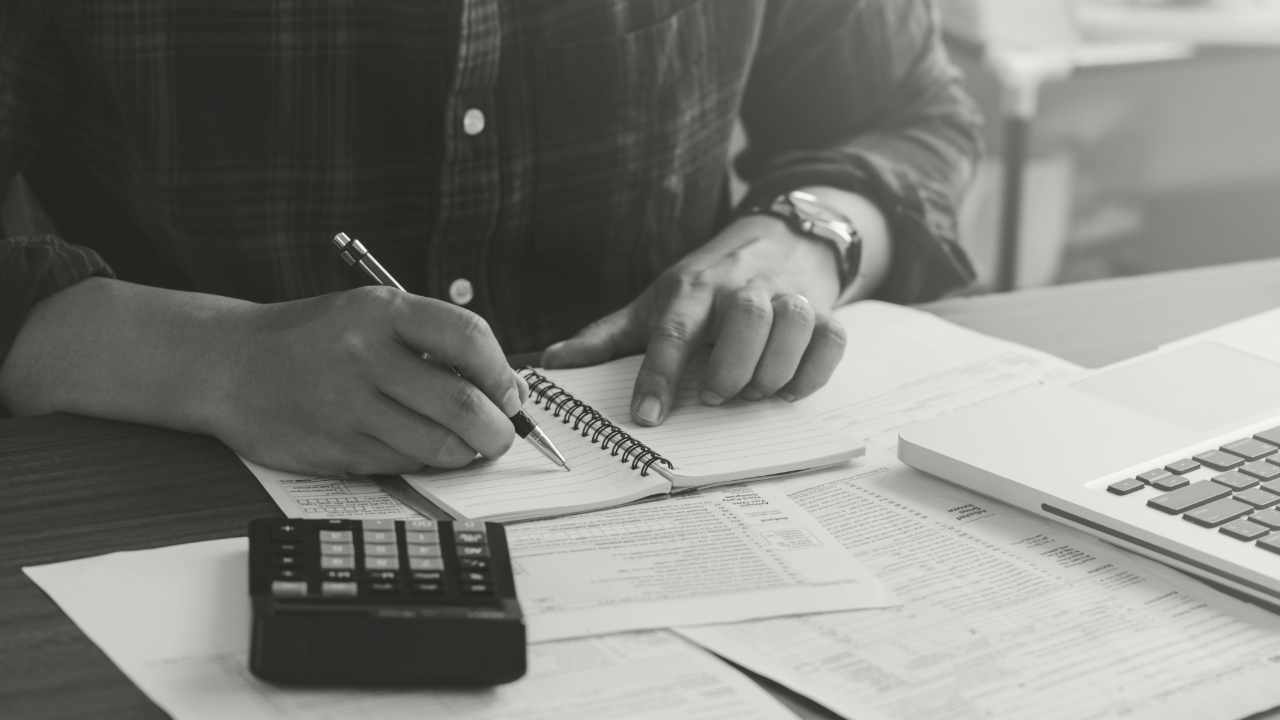 Photo of hands writing on paper with a calculator and computer on a desk.
