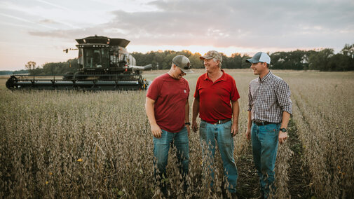 Photo of two generations of farmers standing in a field with a combine in the background.