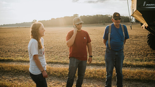 Three people stand outdoors near a field, talking as the sun sets in the background.