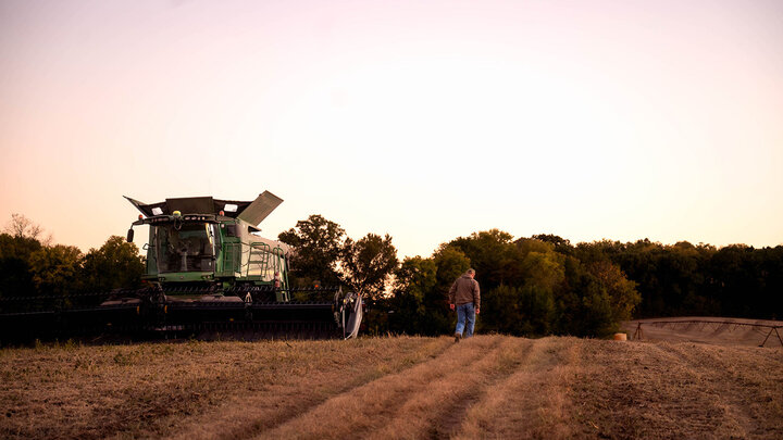 Photo of a farmer walking in a field next to a green combine.