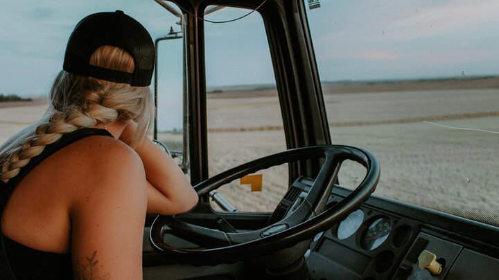 Woman with a blonde braid and black cap sitting in a vehicle, gazing at a vast field landscape.