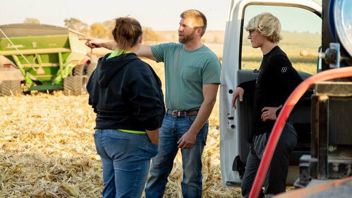 Three people in a field talking near a vehicle; a tractor and harvest equipment visible.