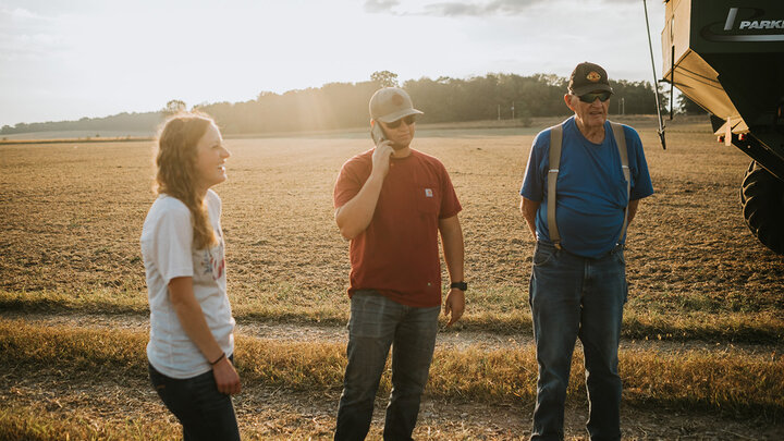 Three people stand outdoors near a field, talking as the sun sets in the background.