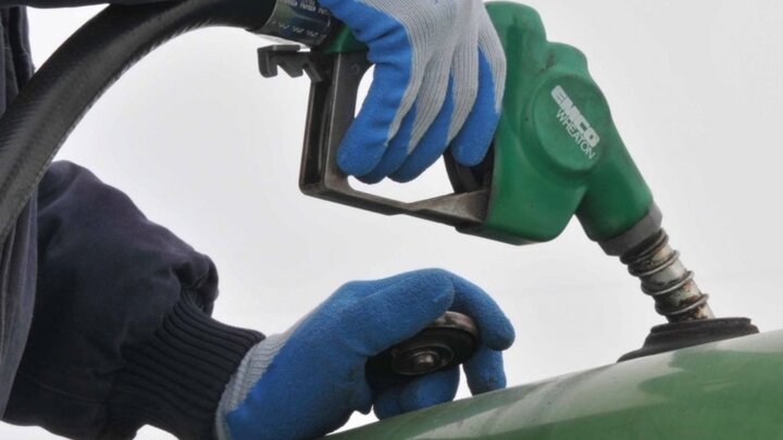 Closeup of gloved hands with a green biodiesel pump inserted into machinery's tank.