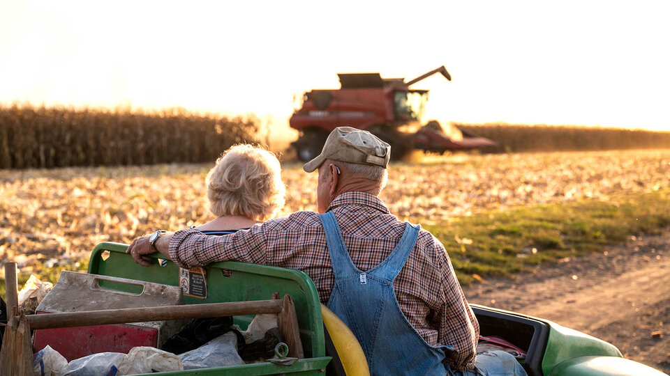 Photo of two senior farmers sitting in a Gator watching a combine harvest corn.