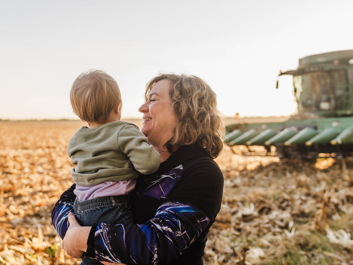 Photo of a woman holding a small child in a field with a combine in the background.