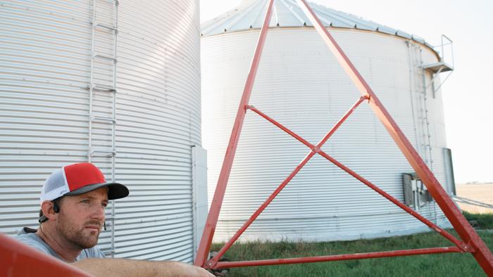 Farmer leaning on equipment in front of grain bins.